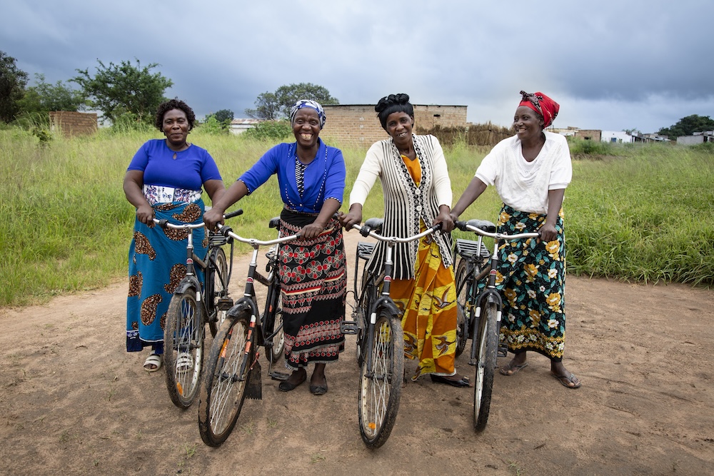 Four smiling women standing outdoors with their bicycles on a dirt path in Luyaba, Zambia