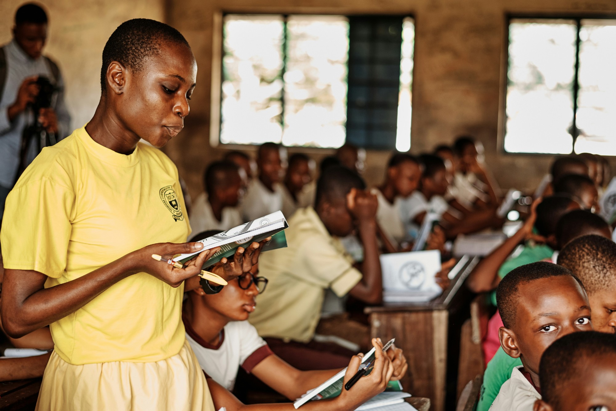 A girl in a yellow school T-shirt stands reading aloud from a booklet in a full classroom, her classmates seated at desks around her