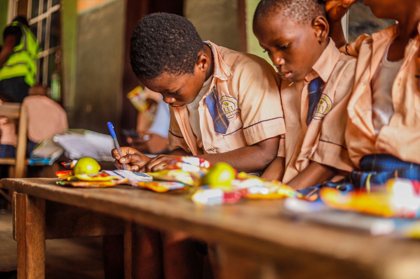 Primary school children in peach uniforms writing at a wooden desk scattered with fruit and snacks